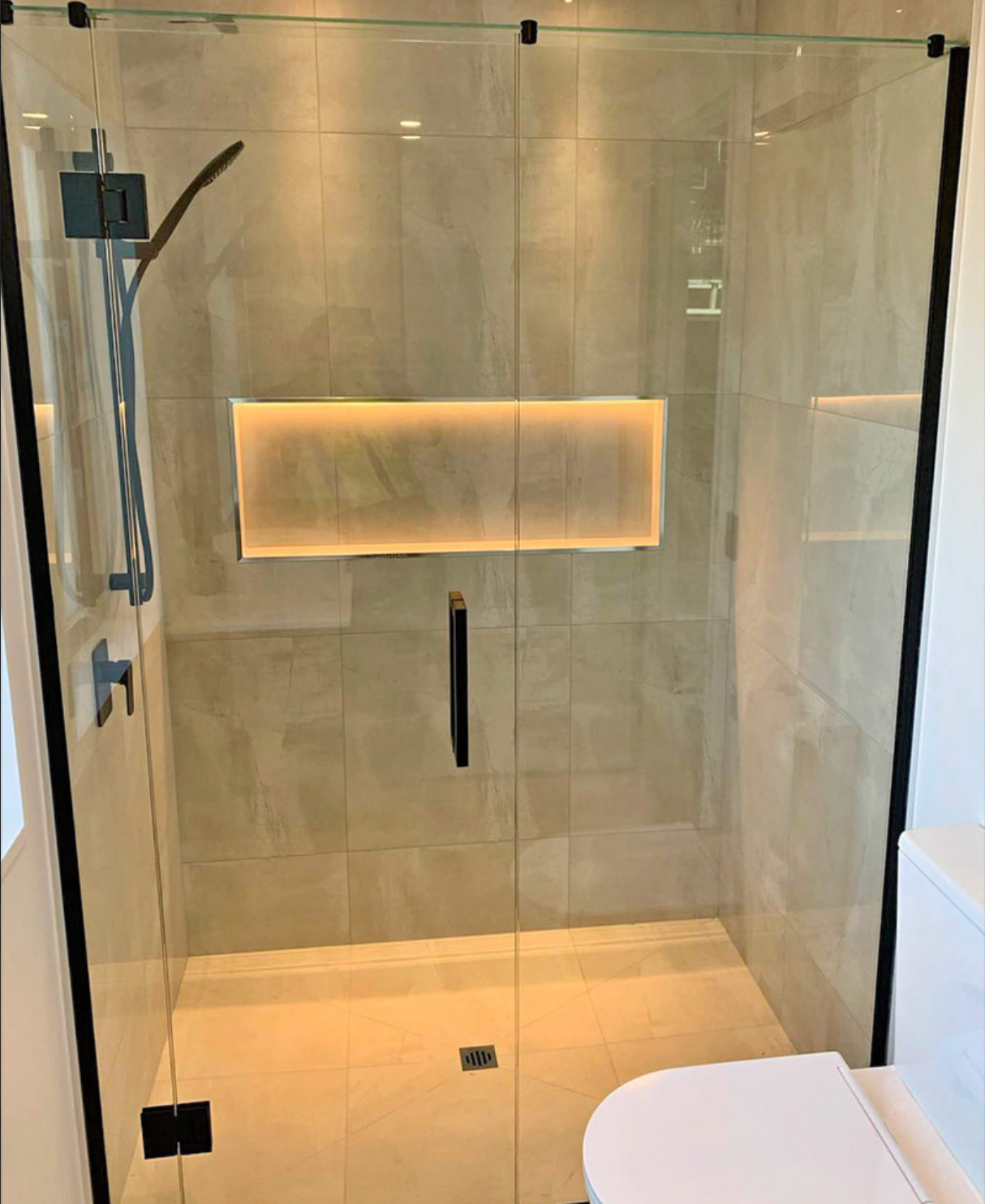 Shower with glass door and black hardware, beige tiled walls, and built-in shelf with lighting.