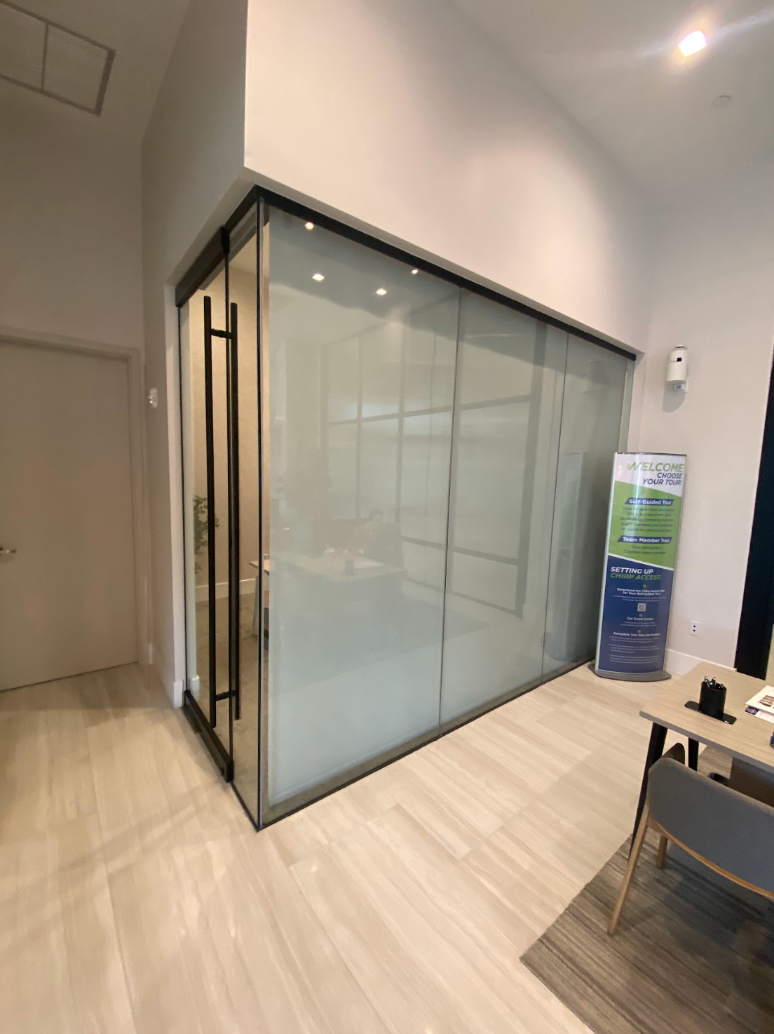 Glass-walled office with frosted glass sliding doors. Light gray interior. A desk with a sign is nearby.