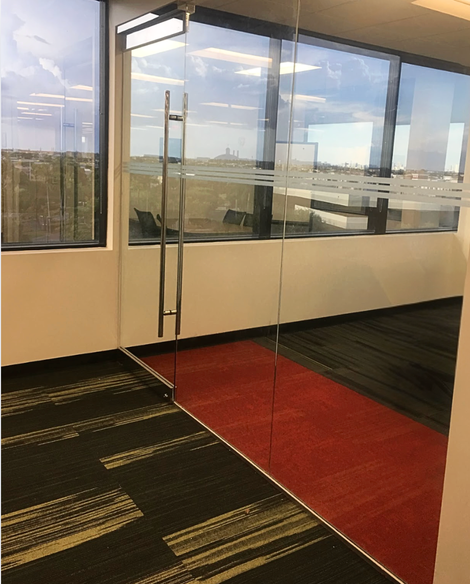 Glass office door with silver handle, red and black patterned carpet, large windows with a view.