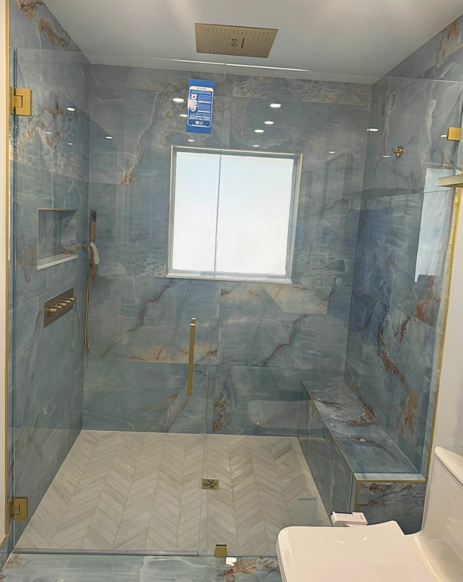 Blue tiled shower with glass doors and gold fixtures, featuring a built-in bench and window.