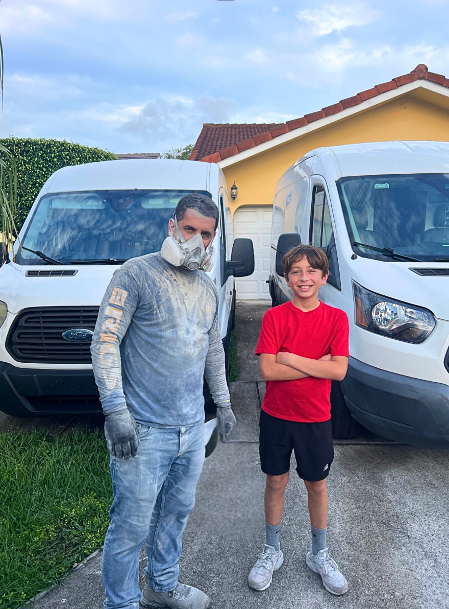 Man covered in dust and boy stand in front of two white vans near a house. Man wears a mask.