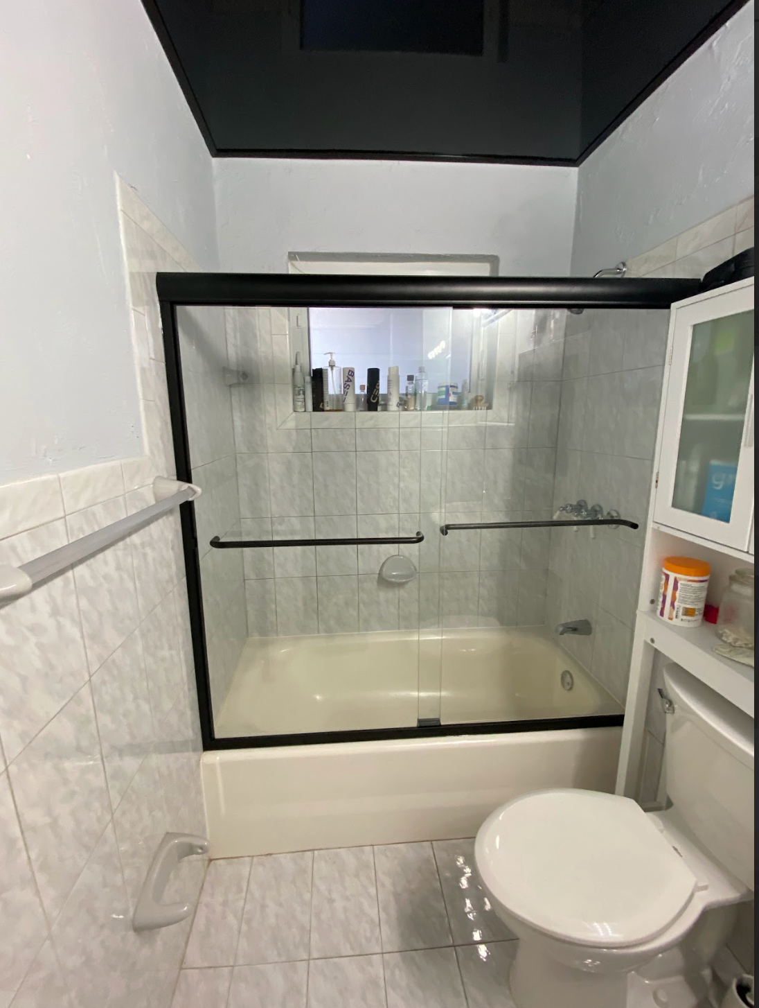 Bathroom with white tiled walls, black shower frame, toilet, and white cabinet.