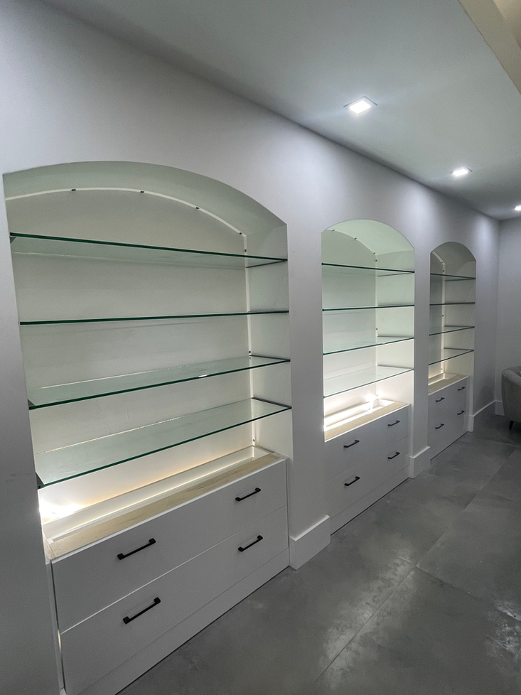 Built-in white shelves with curved tops, glass shelves, and drawers, illuminated by lights, against a gray wall.