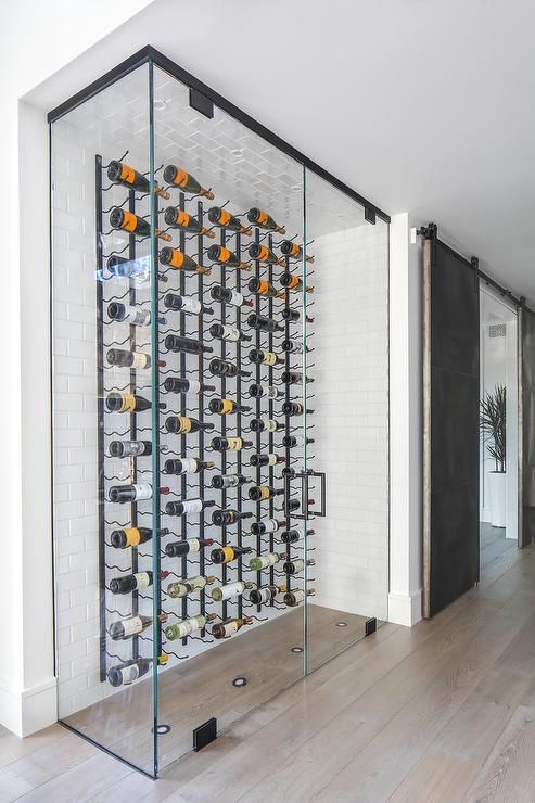 Glass-enclosed wine cellar with rows of bottles, black metal rack, and sliding barn door in a modern hallway.