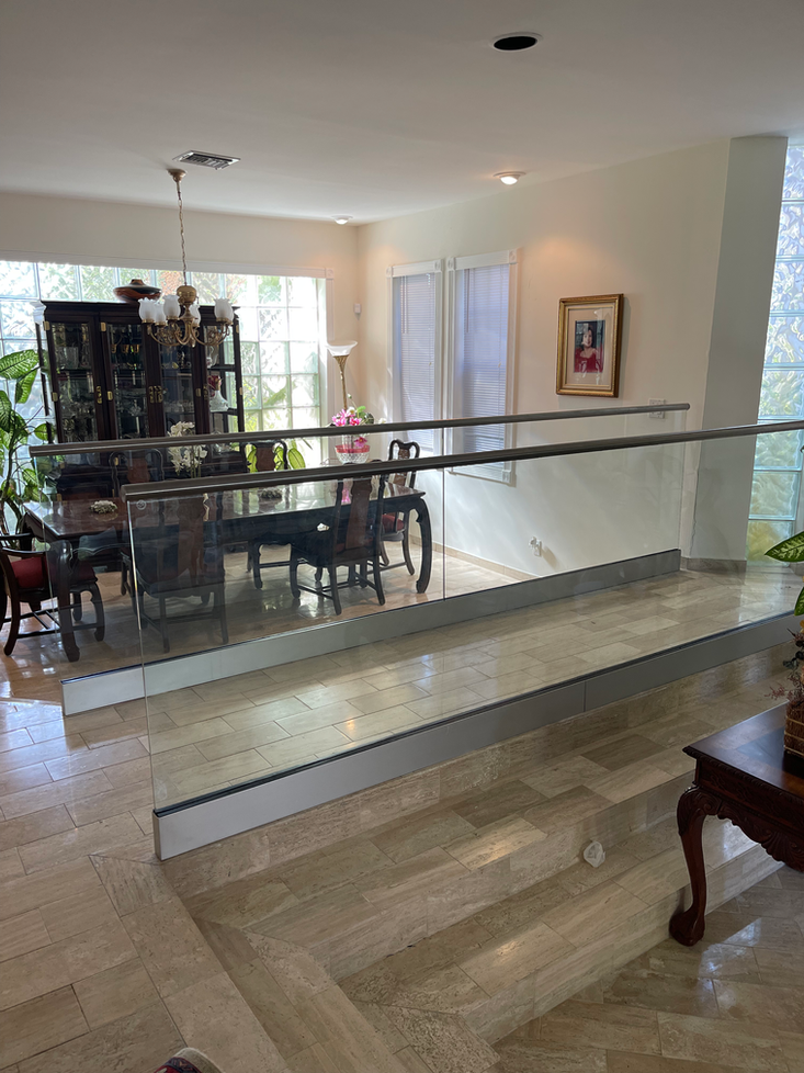 Dining room with dark table, chairs, and glass railing; cream-colored flooring, windows, and decorative items.