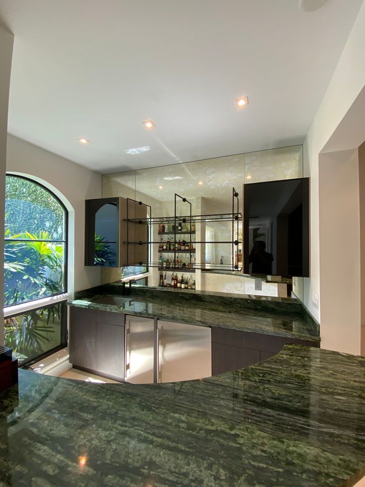 Bar with green countertop, cabinets, and open shelving against a mirrored wall, near a large arched window.