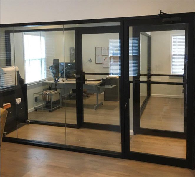 Glass-walled office with black frames and door. Inside, a desk and computer are visible.