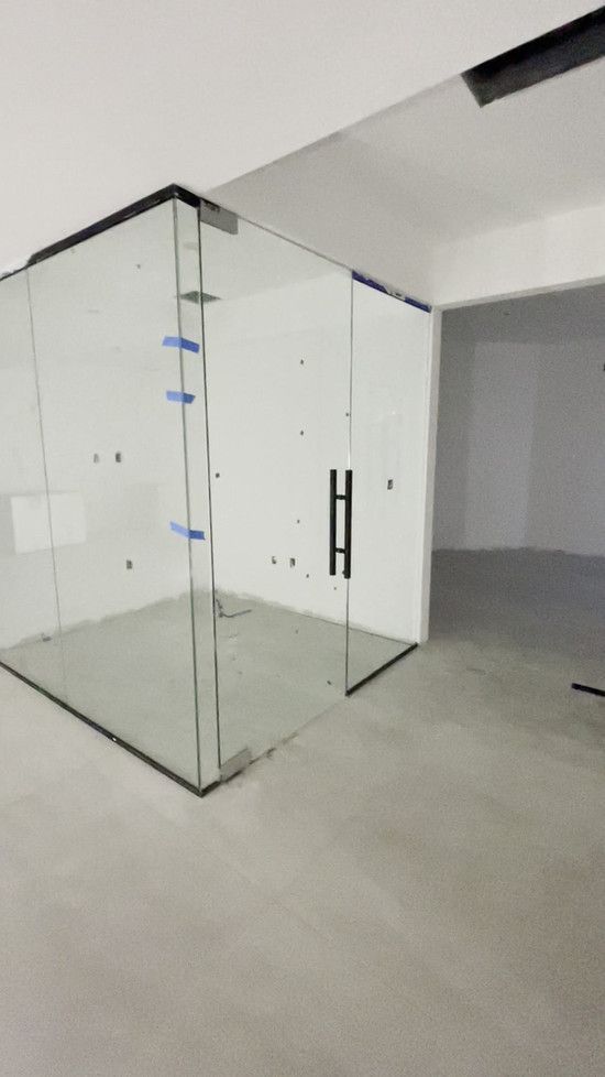 Glass enclosed room with black door handle in a room with white walls and gray floor.