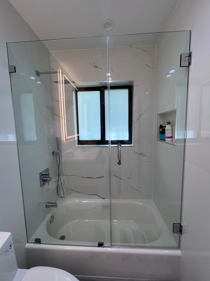 Glass shower enclosure over a white bathtub. The enclosure features a door and is set against white marble tiles.