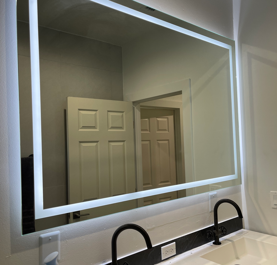 Lighted mirror above a bathroom vanity with black faucets, reflecting a doorway and closed door.