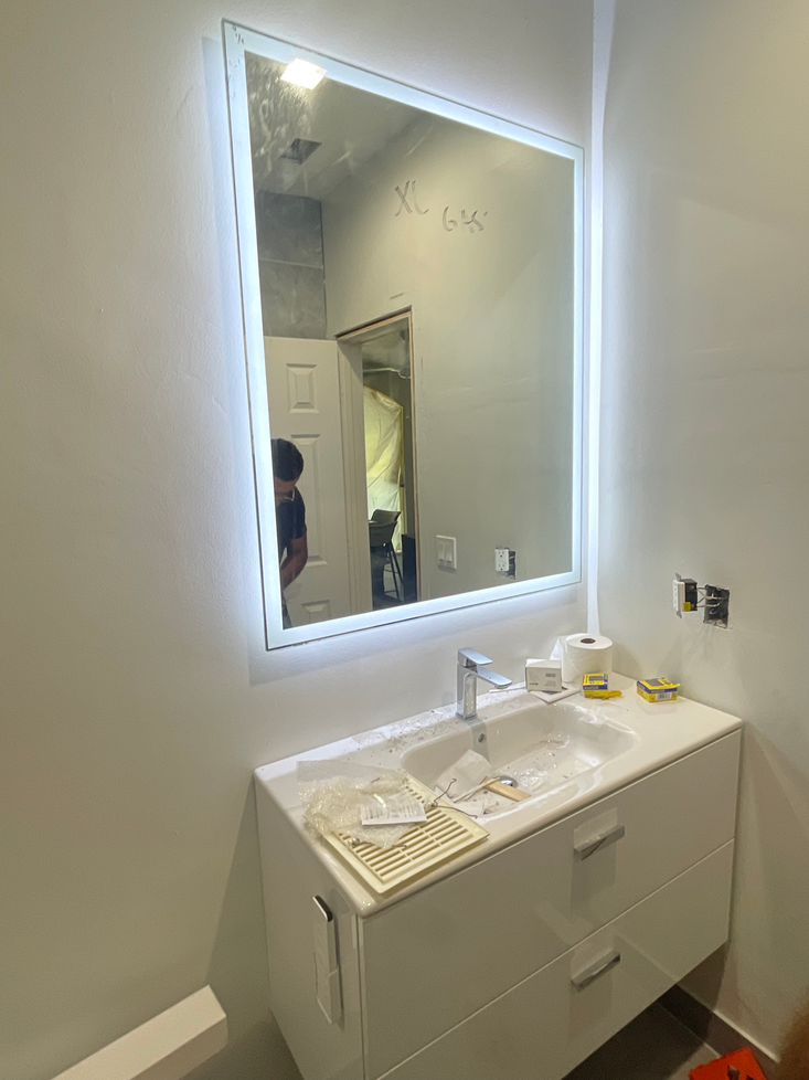 Bathroom with a lit-up mirror above a white vanity and sink. A person is reflected in the mirror.