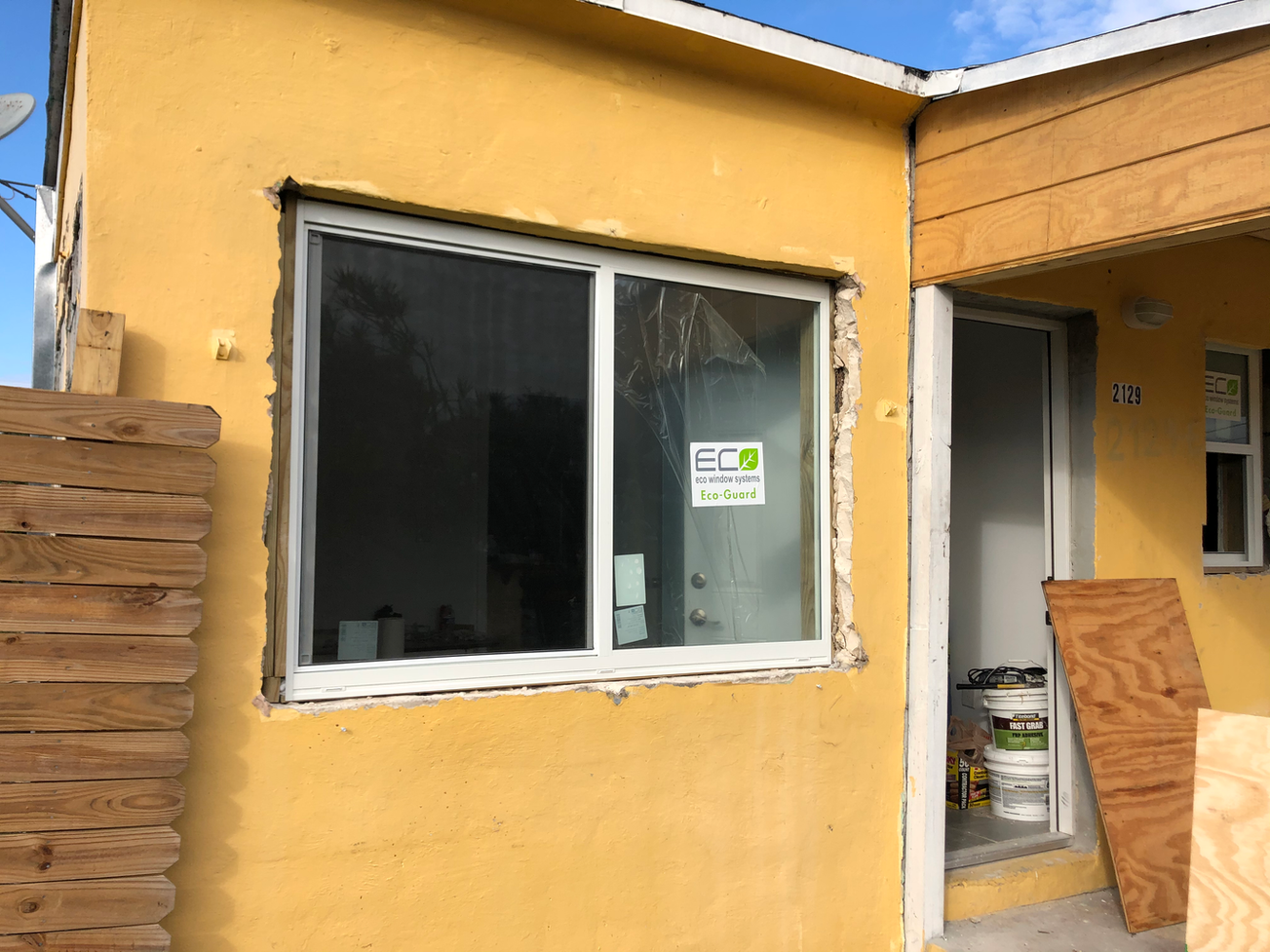 Yellow stucco building with a white-framed sliding window. A plywood board rests near a door.