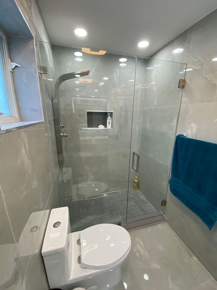Modern bathroom with a glass shower, toilet, and gray tile walls. Teal towel hangs on the shower door.