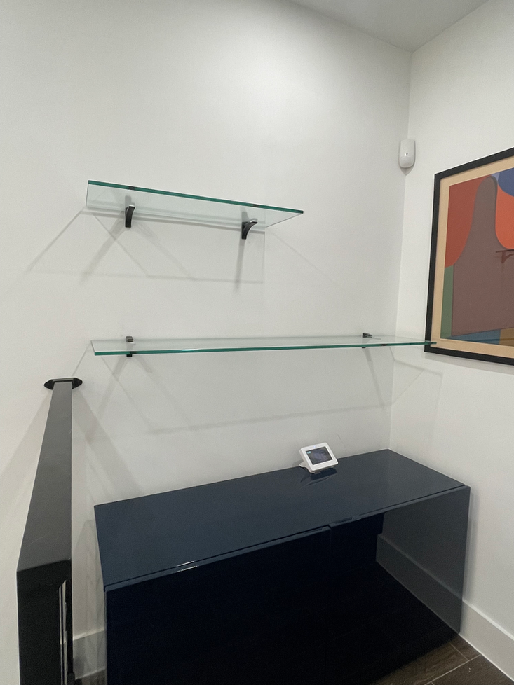 Two glass shelves on white wall, above a dark blue cabinet. A framed abstract painting and security device are also visible.