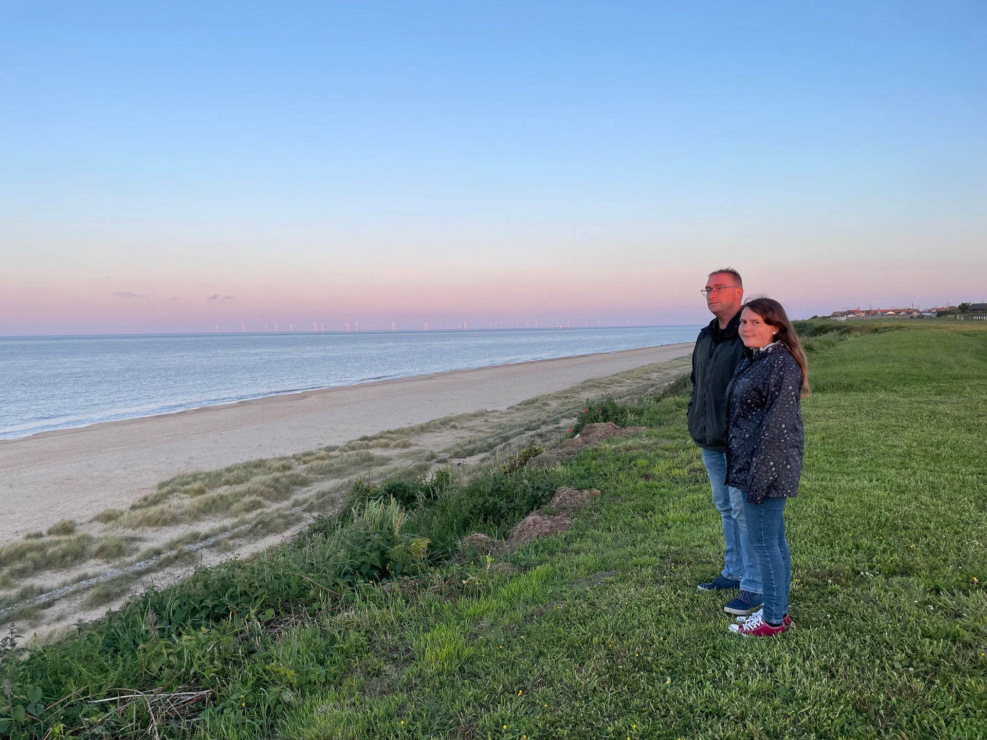 A man and a woman are standing on a grassy hill overlooking the ocean.