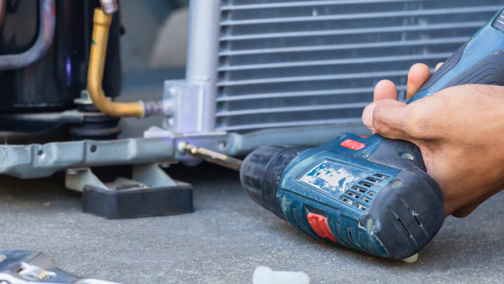 Hand using a blue power drill to loosen a car bumper near the ground