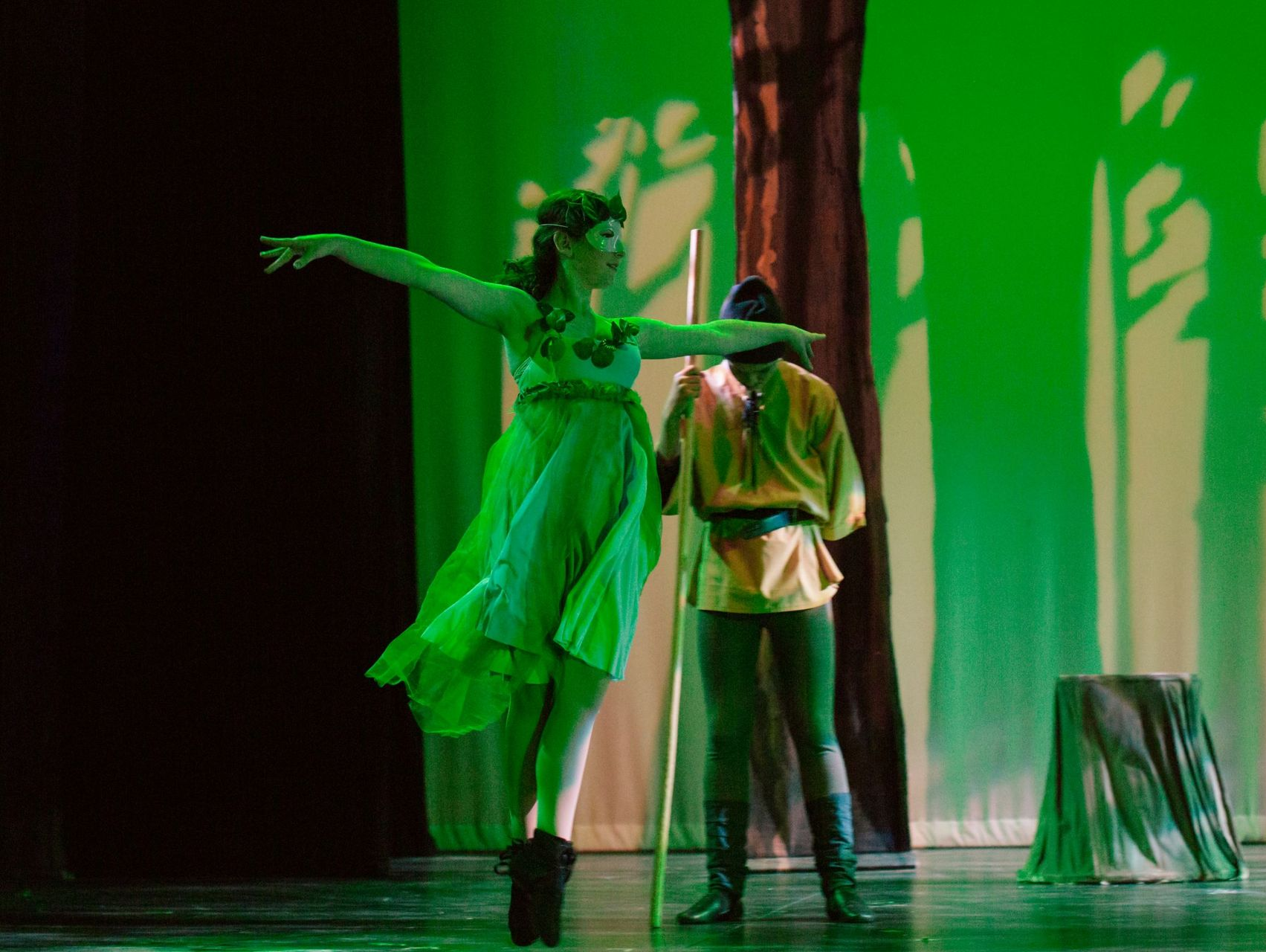 Two performers on stage, one in a bright green dress and one in a yellow tunic, lit by green spotlights