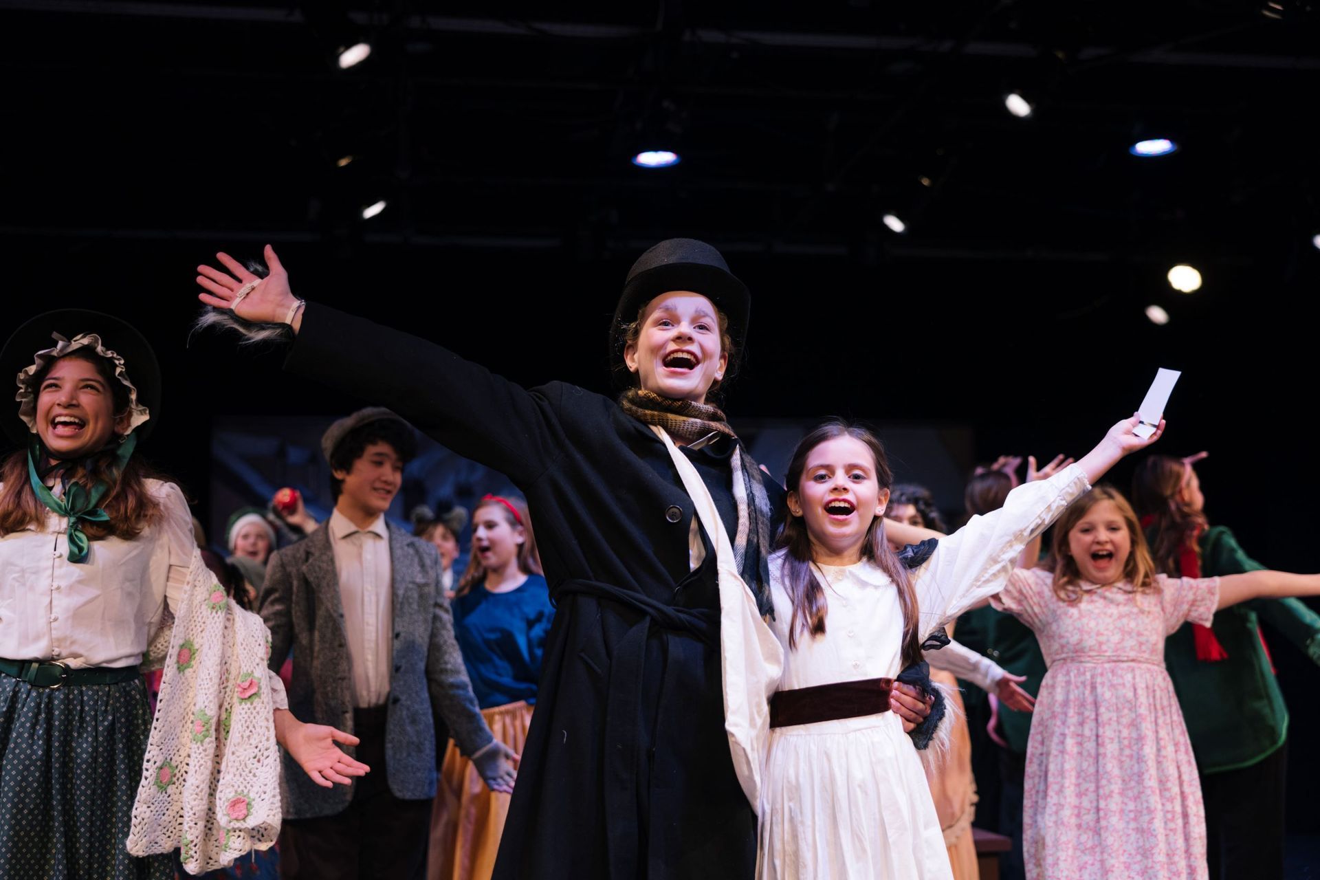 Performers on stage with arms raised, smiling under bright lights in a theatrical finale.