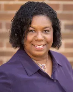 Portrait of a smiling person in a purple blouse against a brick background
