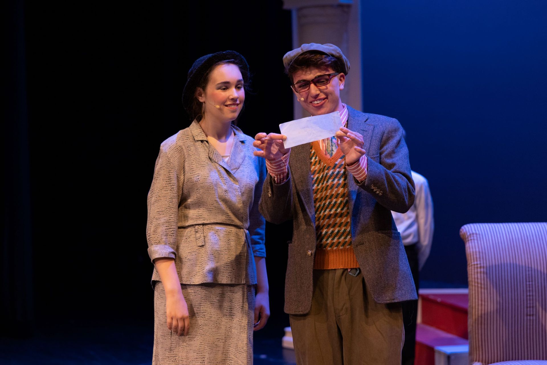 Two actors onstage, one holding a letter and smiling beside the other under stage lights.