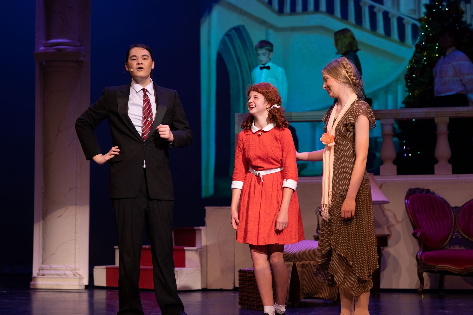 Three actors onstage in colorful costumes, standing before a theater set with columns and blue lighting