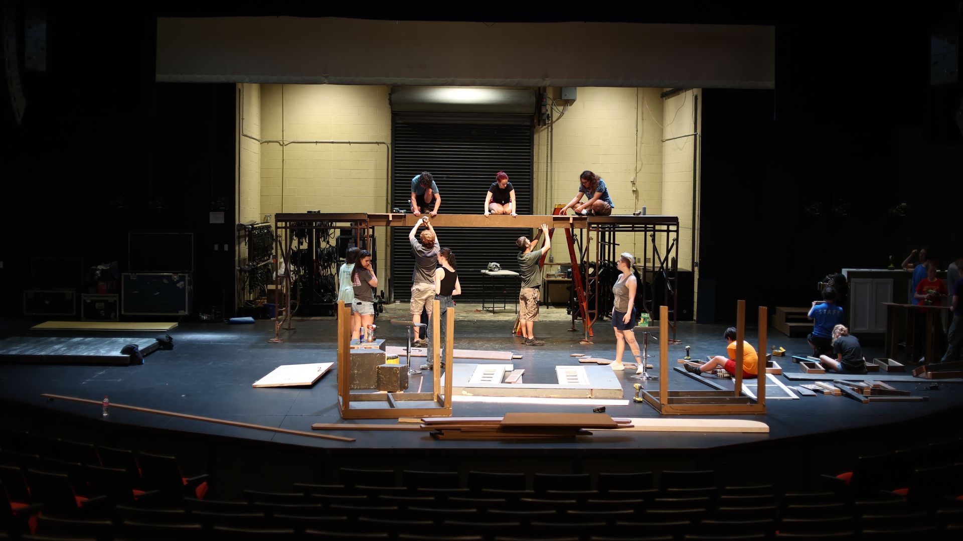 Theater stage with several people and a wooden set piece under construction under stage lights