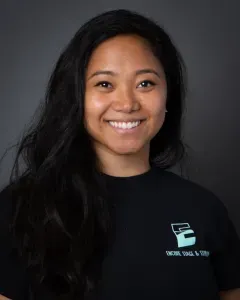 Smiling person with long dark hair wearing a black shirt against a gray background