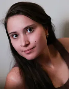 Portrait of a woman with long dark hair and a slight smile, leaning against a light wall.
