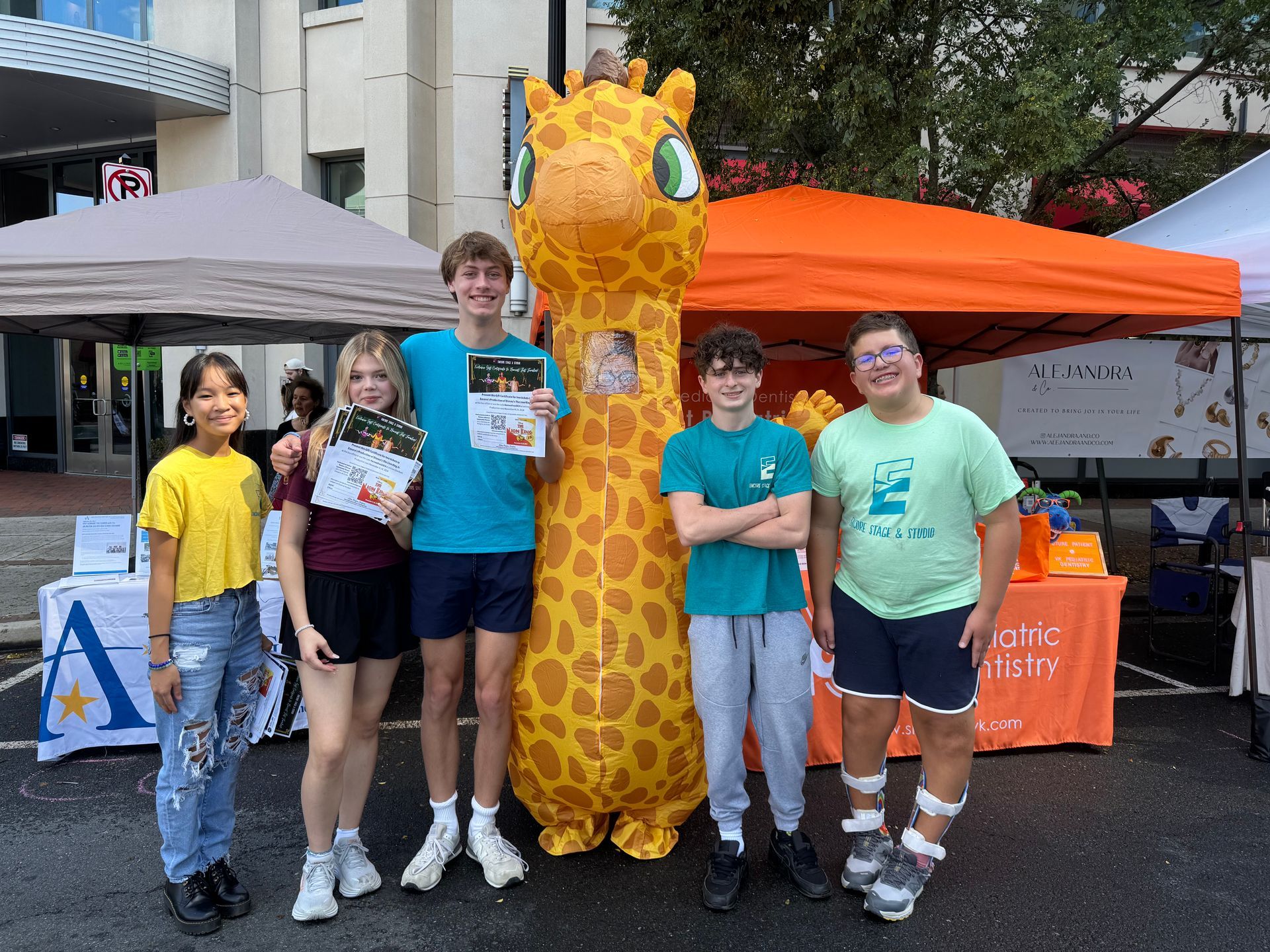 Five people pose with a giraffe mascot in front of orange and gray event tents.