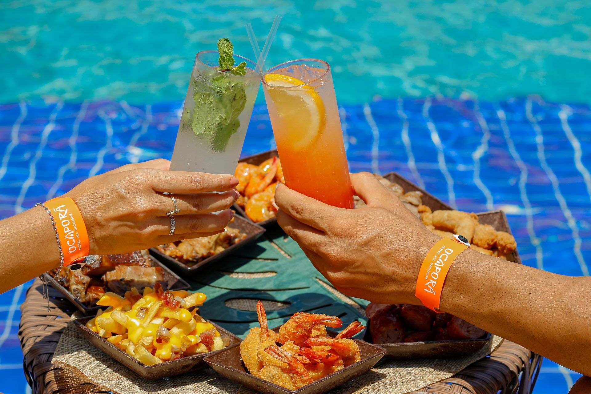 Um casal está brindando com bebidas e comidas ao lado de uma piscina.