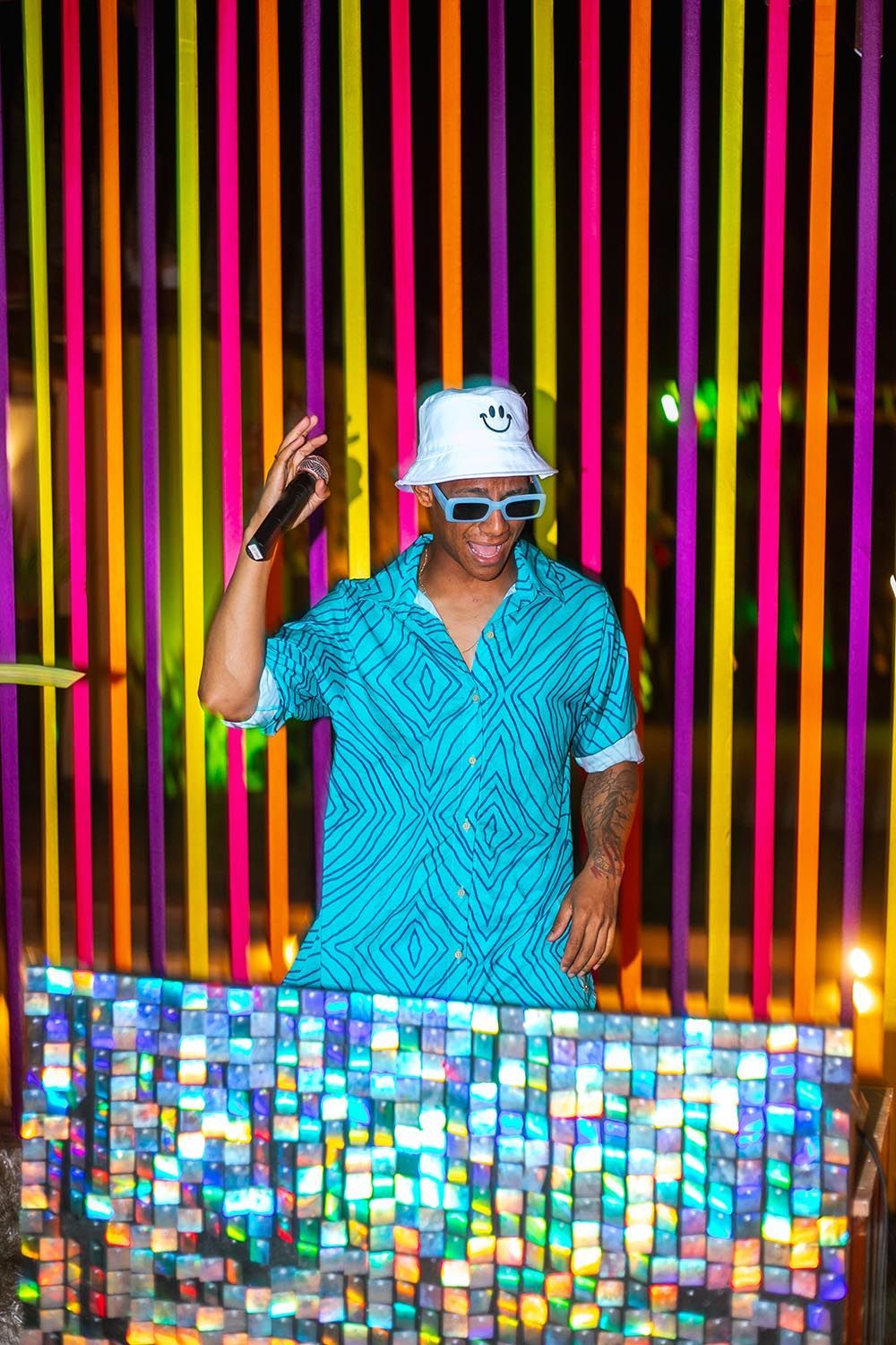 Um homem de camisa azul e chapéu branco está parado em frente a uma parede colorida.