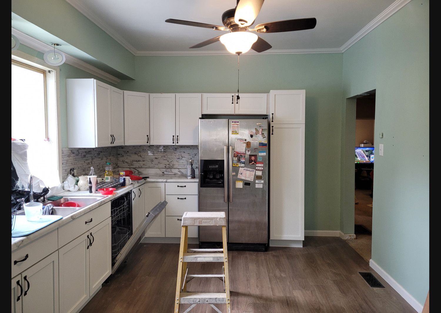 Bright kitchen with white cabinets, stainless fridge, ceiling fan, and a small step ladder in the center.