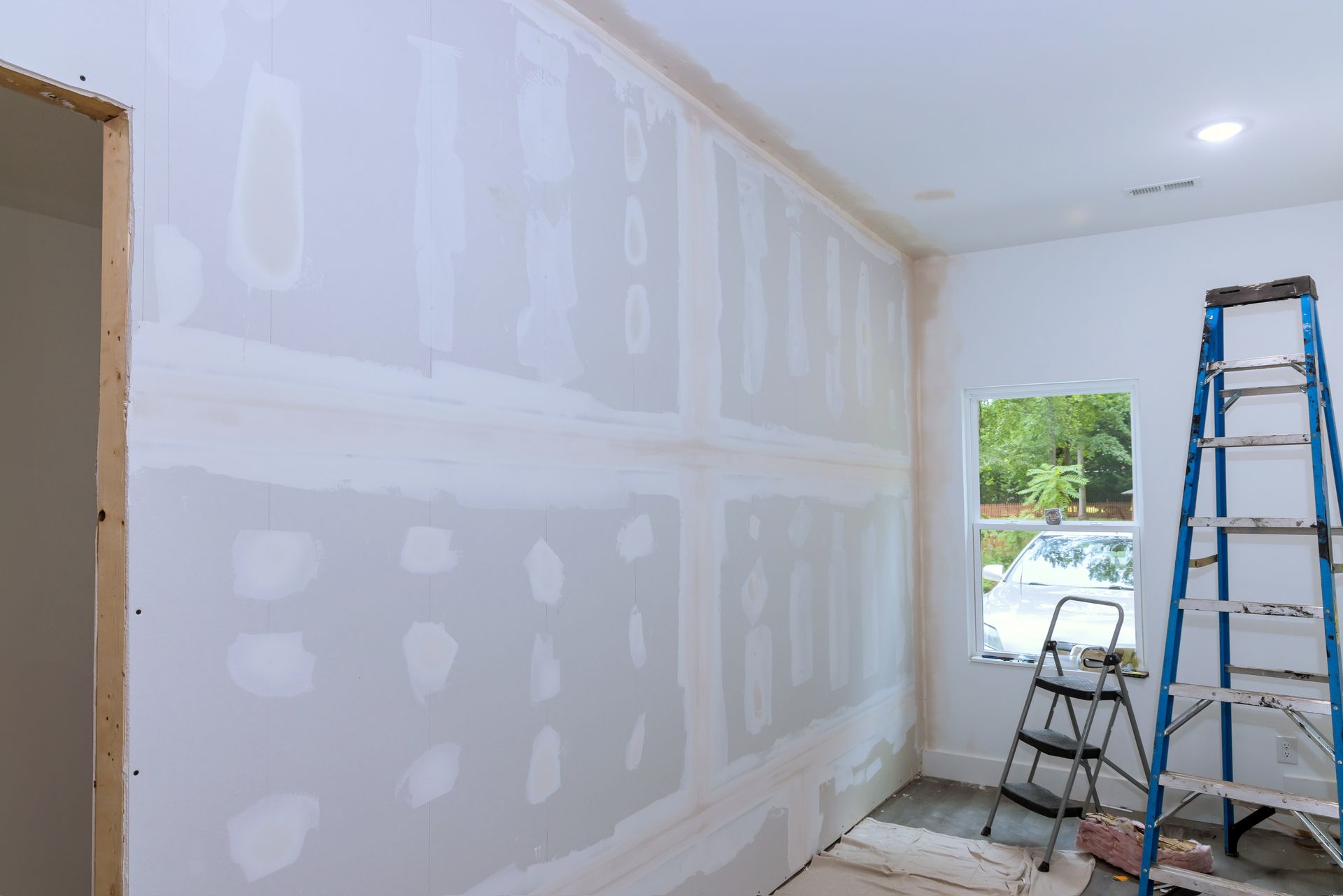 Room under renovation with taped drywall patches and ladders near a window and open doorway