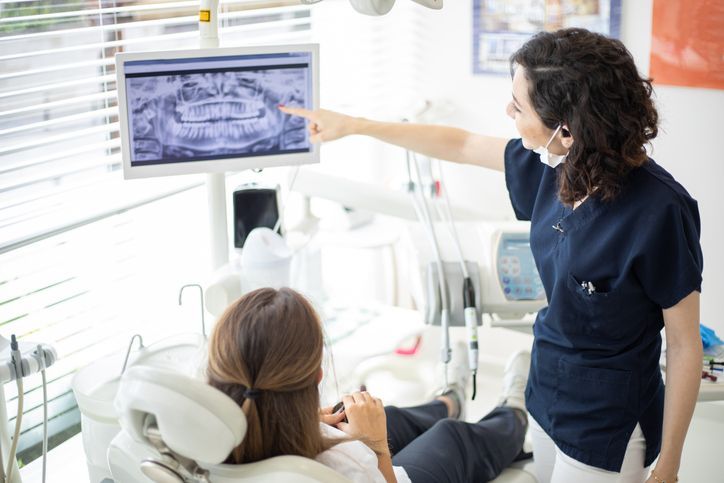 Dentist points to an X-ray on a monitor while a patient reclines in the dental chair.