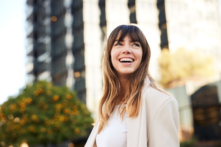 Smiling woman in a white blazer standing outdoors near blurred buildings and orange trees