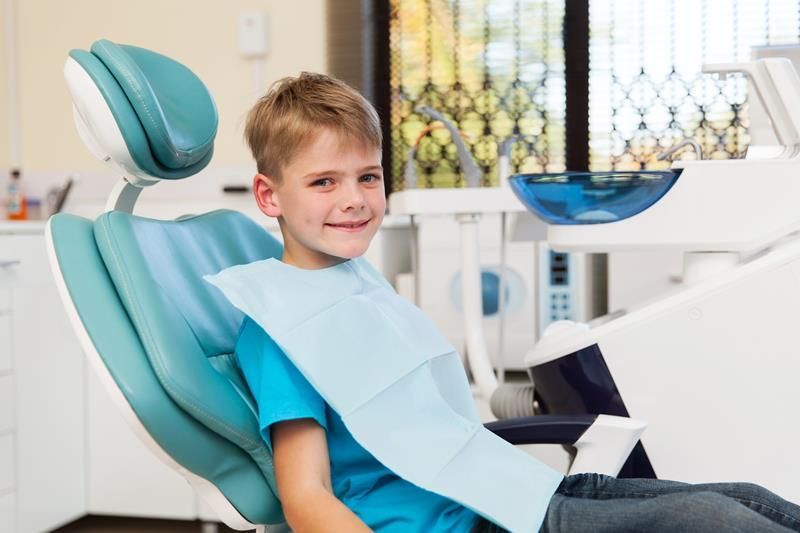 Smiling child in a turquoise dental chair wearing a bib at a dentist’s office