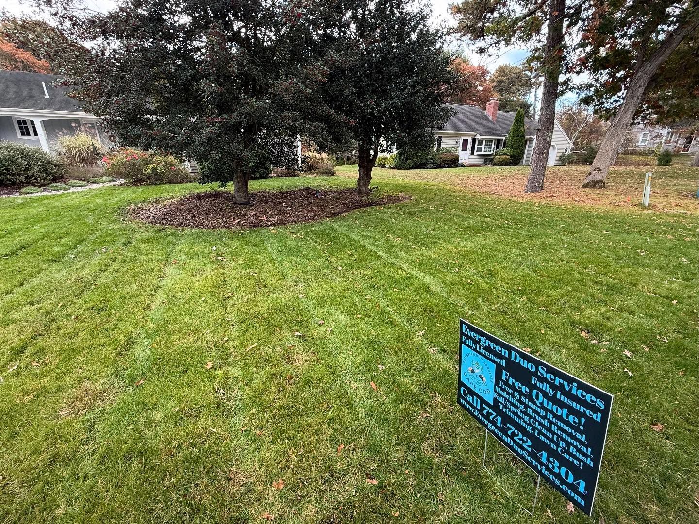 Green lawn with trees, houses in the background, a sign in the foreground with text and phone number.