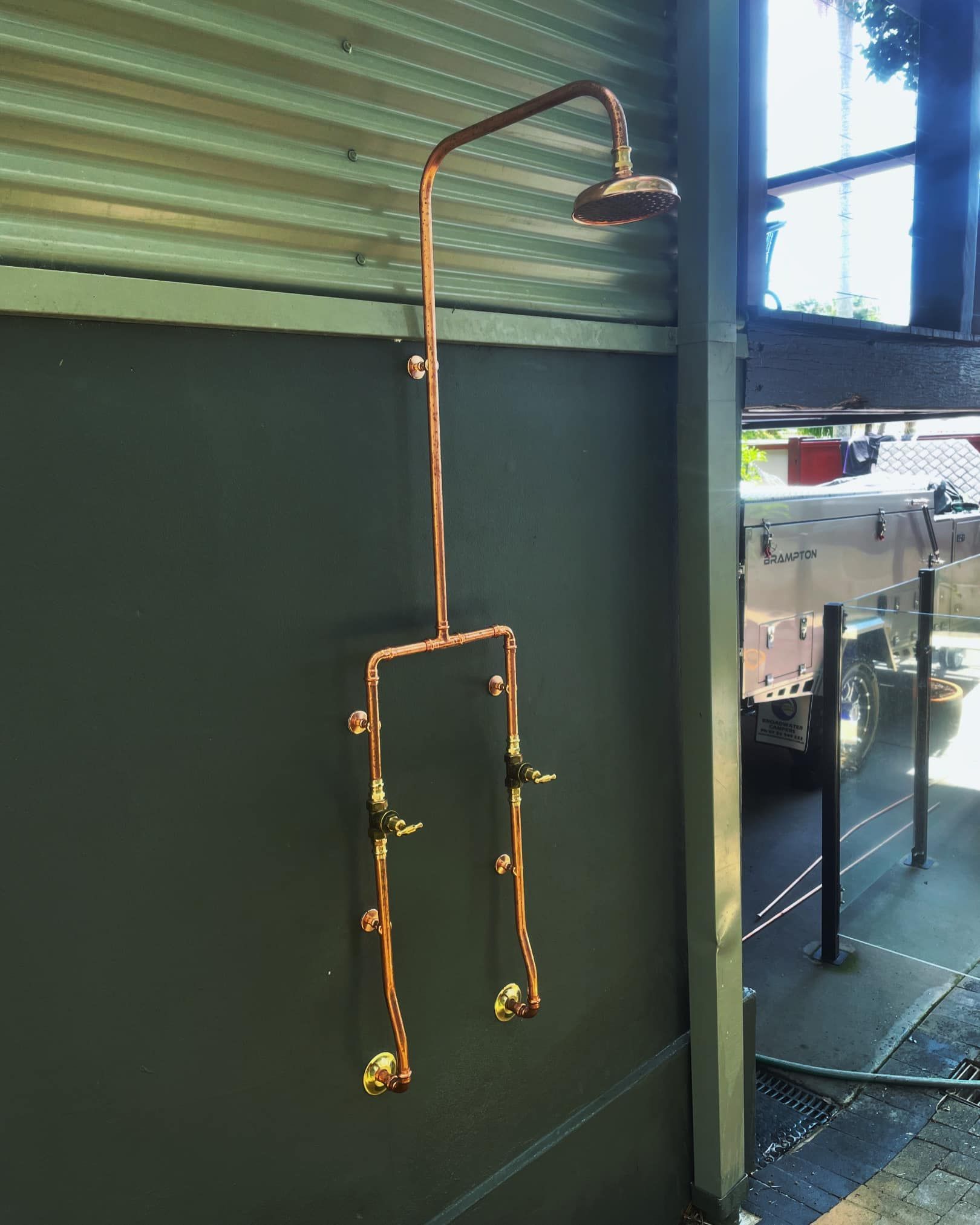 Copper shower fixture mounted on a green wall, with a curved shower arm and exposed pipes. — New Gen Plumbing and Gas in Banora Point, NSW