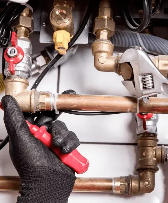Gloved hands using red-handled pliers to grip a copper pipe under a sink, with brass fittings and valves. — New Gen Plumbing and Gas in Coolangatta, NSW