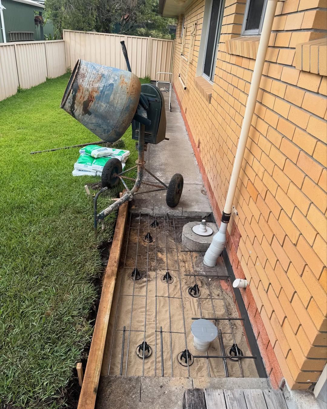 Concrete side yard beside brick house with wheelbarrow, cement mixer, and tools along a fence — New Gen Plumbing and Gas in Murwillumbah, NSW