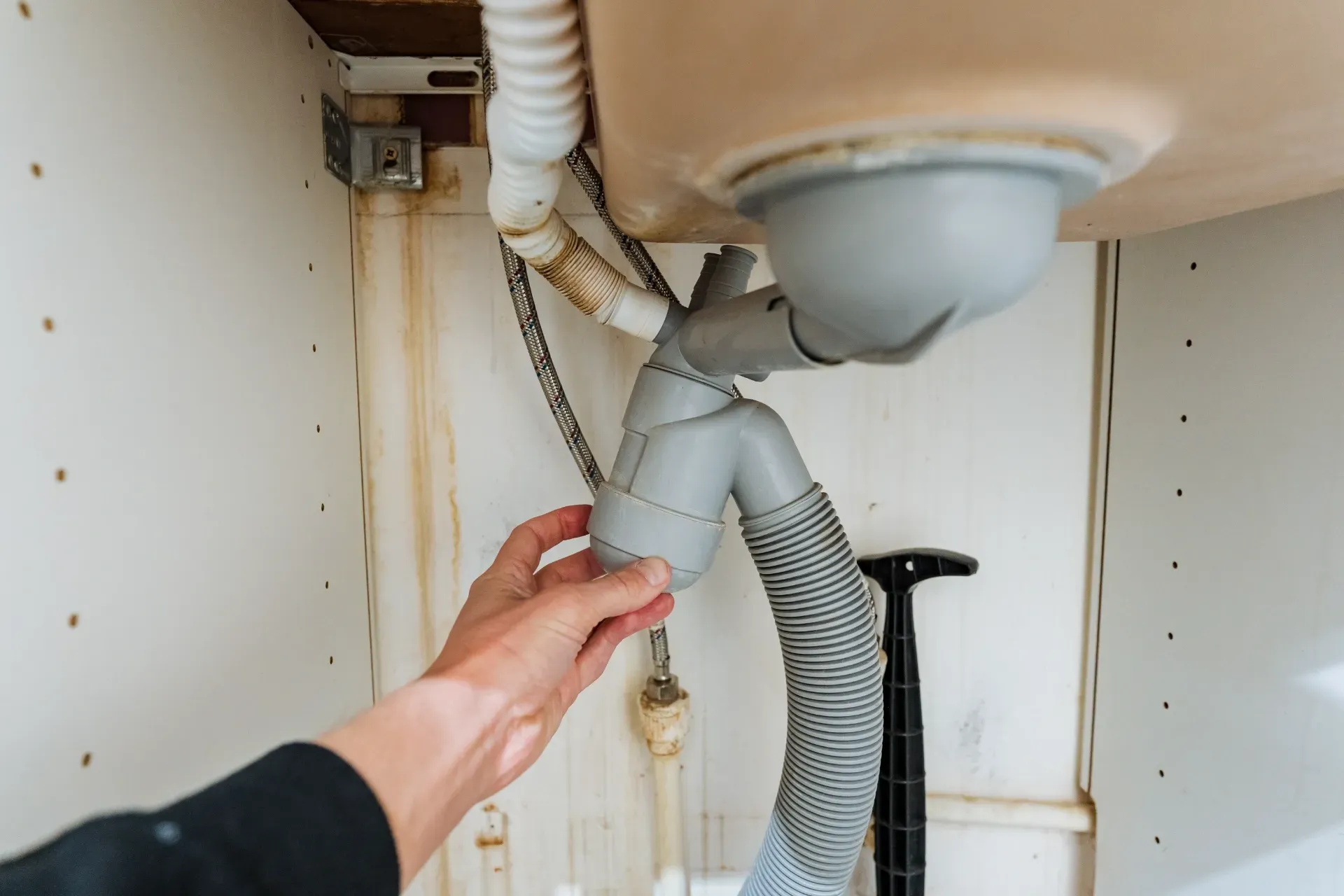Hand reaching under a sink to adjust a gray drain hose and pipe connections — New Gen Plumbing and Gas in Banora Point, NSW