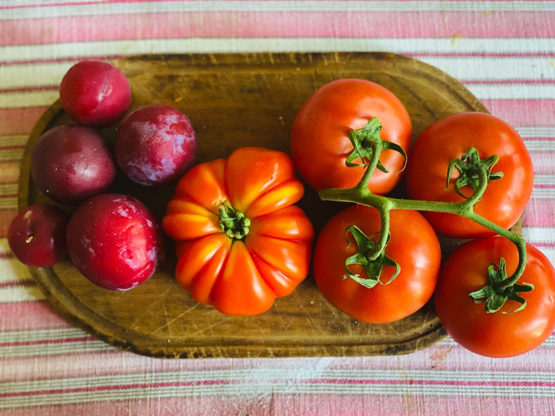 Plums and tomatoes on a wooden cutting board, resting on a striped tablecloth.
