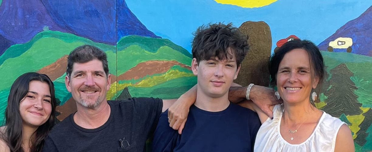 Family of four smiling in front of a colorful mural of a landscape scene.