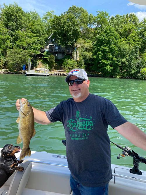 Man on a boat holding up a fish on a green lake with trees and a house in the background