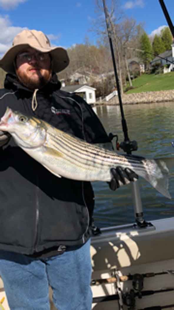 Man holding a striped fish on a boat, wearing a hat and jacket on a lake.