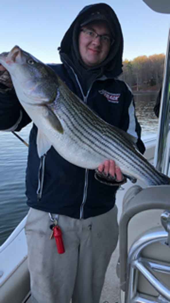 Person holding a large striped fish on a boat in the water, wearing a hooded jacket.