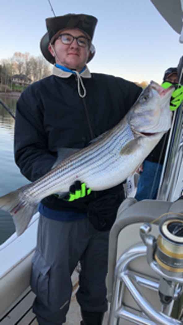 Person holding a large striped fish on a boat, wearing a dark jacket and hat.