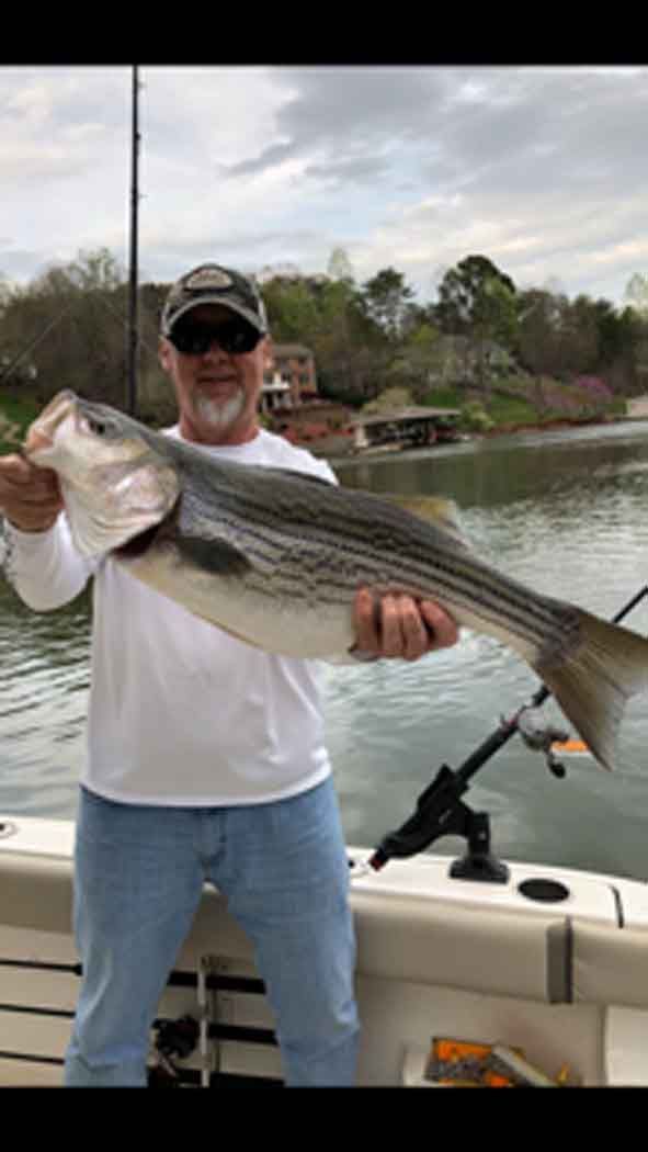 Man on a boat holding a large striped fish on a calm lake under cloudy skies