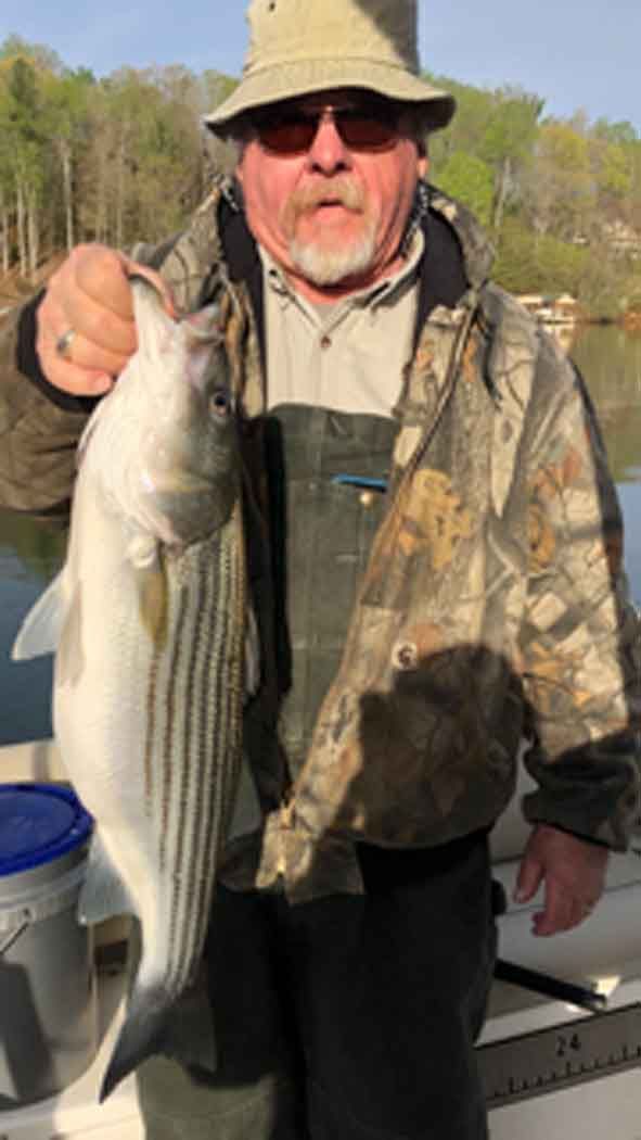 Man holding a large striped fish on a boat, wearing a hat and camo jacket, with water and trees behind him