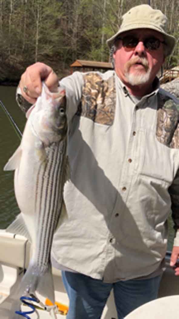 Man holding a large striped fish on a boat, wearing a tan shirt, hat, and sunglasses outdoors near water